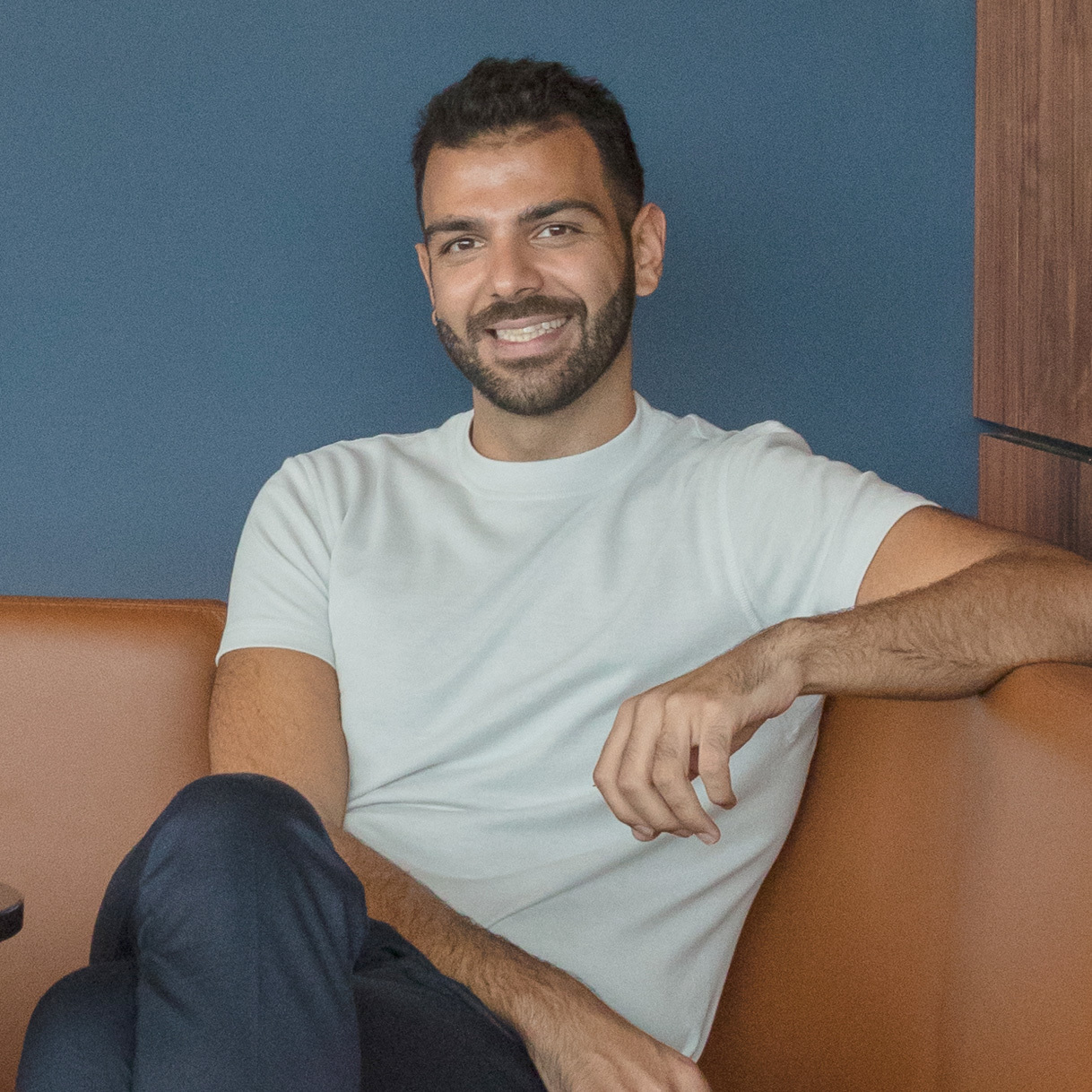 Smiling man with dark hair and beard wearing light green t-shirt, sitting on orange sofa against blue wall with wooden trim.