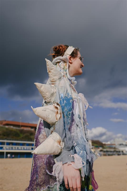 A person wearing a pollution-inspired dress on Bournemouth beach, blue, green and white with patterns and white spines.