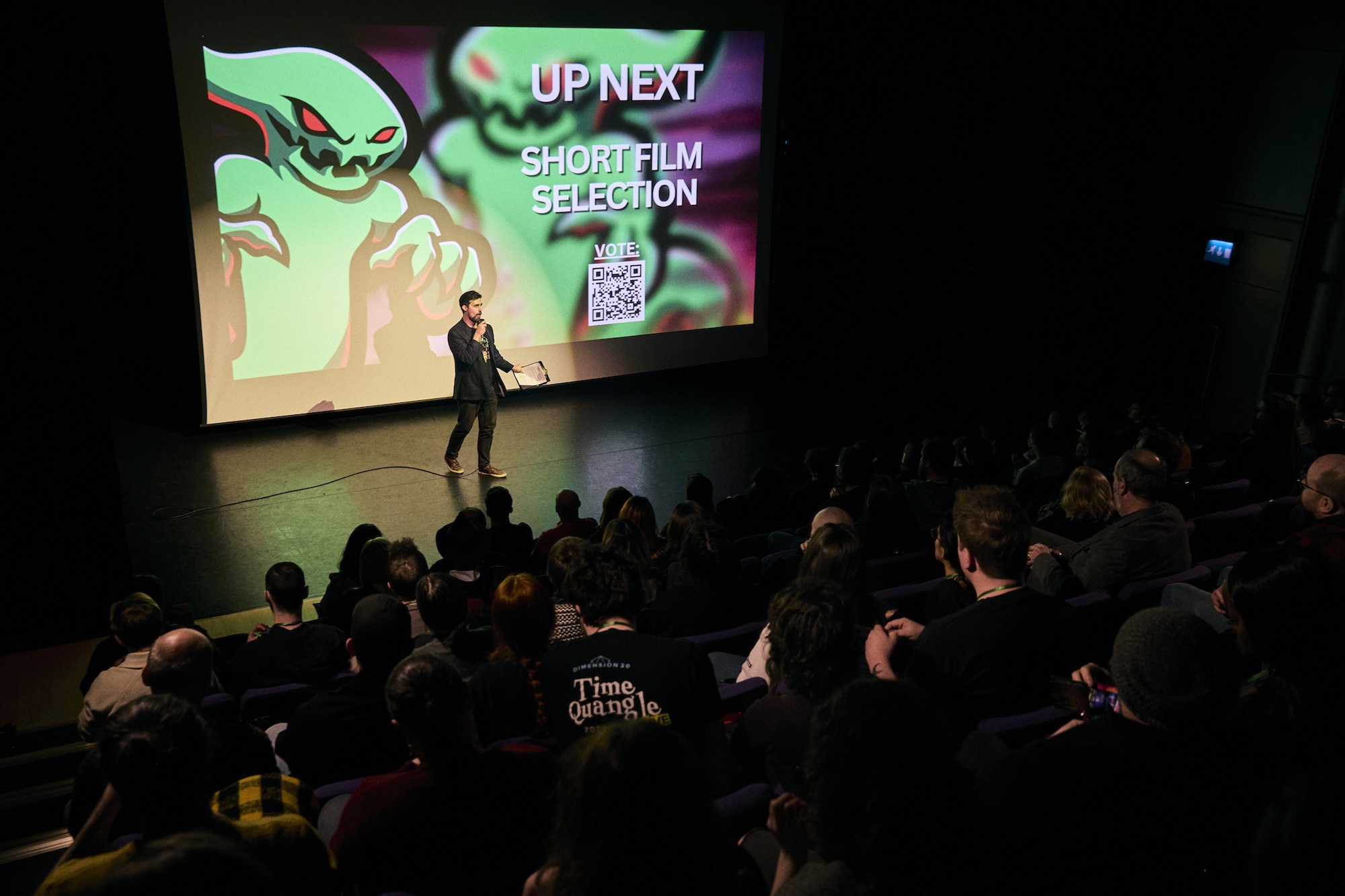 R Nikolov stands on a dark stage in front of a large screen reading 'UP NEXT SHORT FILM SELECTION', with an audience seated in rows below.