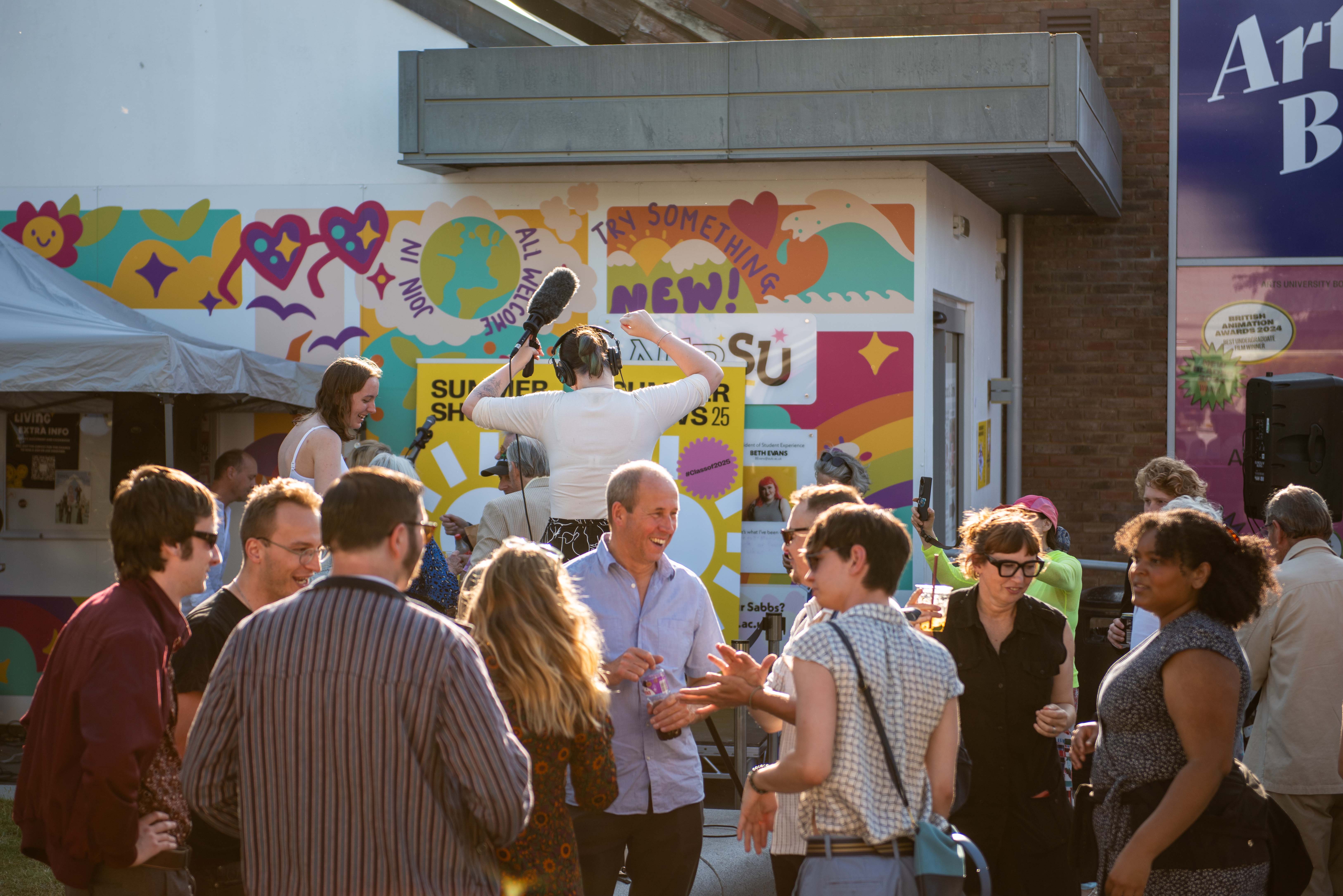 People talking and person with headphones and boom mic dancing on AUB campus in the sun, colourful AUBSU mural in the background.