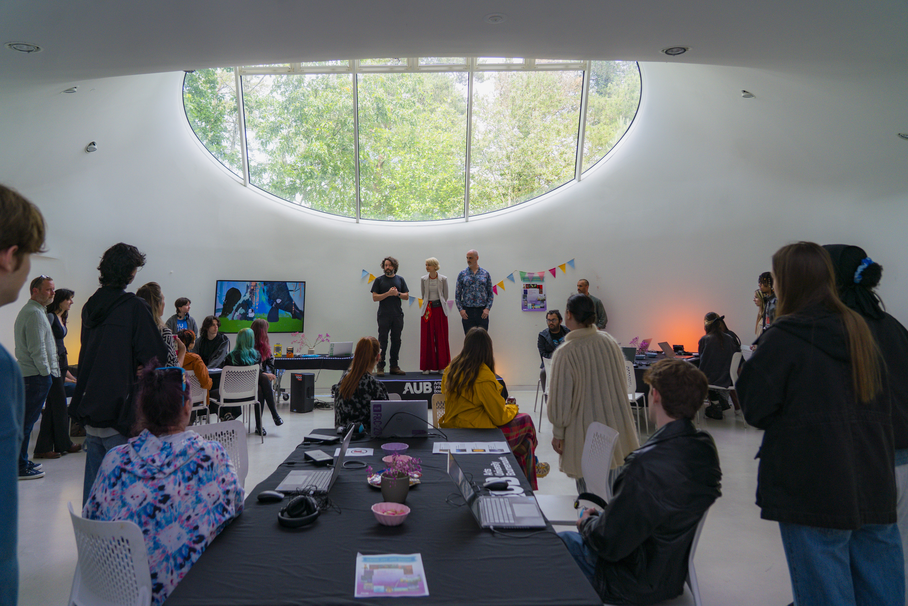 People in AUB Drawing Studio, table, display and orange lighting with three course leaders stood on an AUB podium.