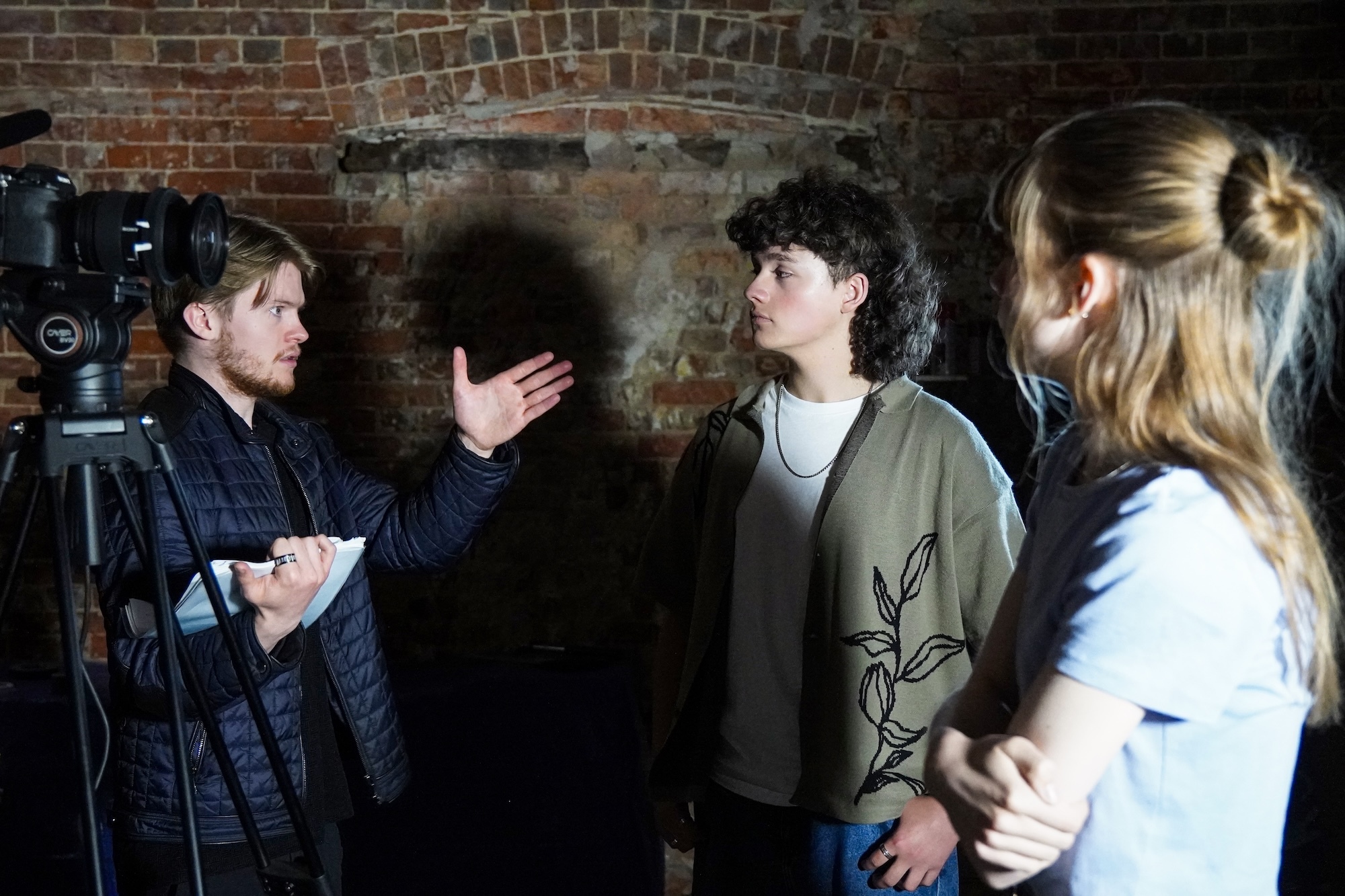 J Rigby with film crew members in conversation against brick wall backdrop with filming equipment visible on left side.