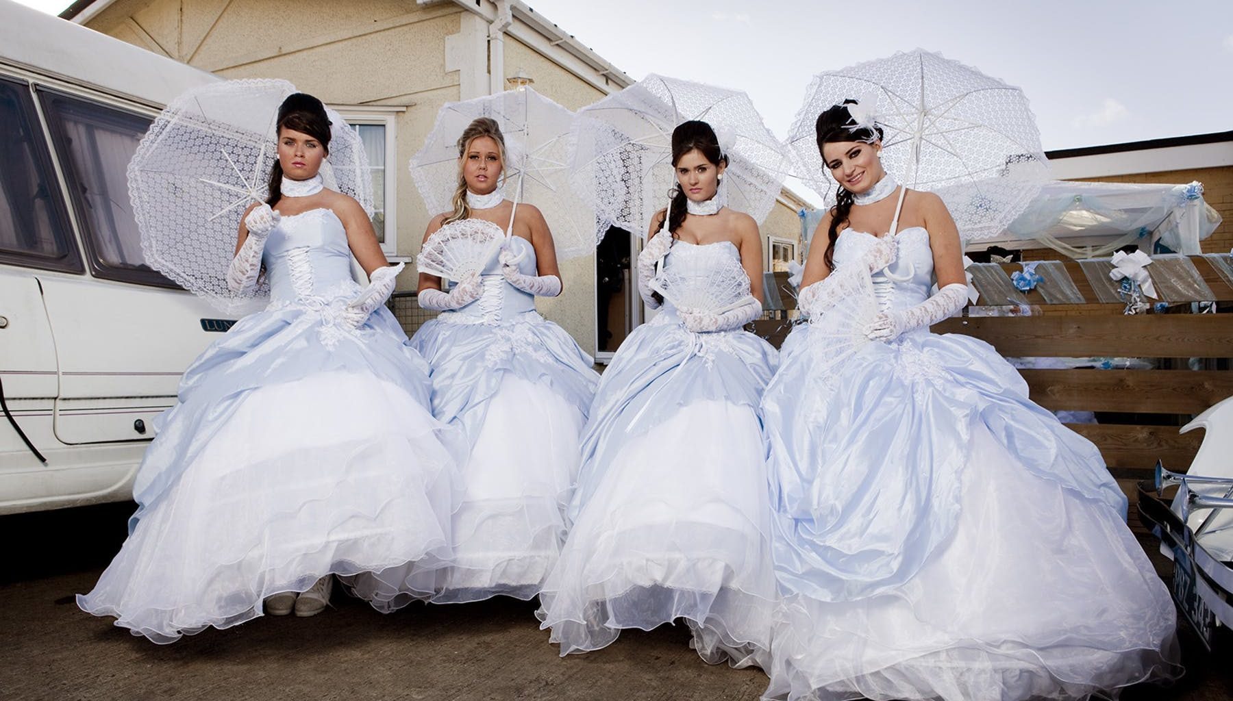 Four women in wedding dresses and parasols pose for a photo next to a caravan.
