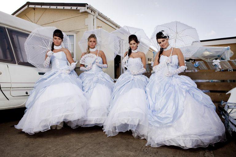 Four people in identical wedding dresses pose for a photo next to a caravan holding parasols.