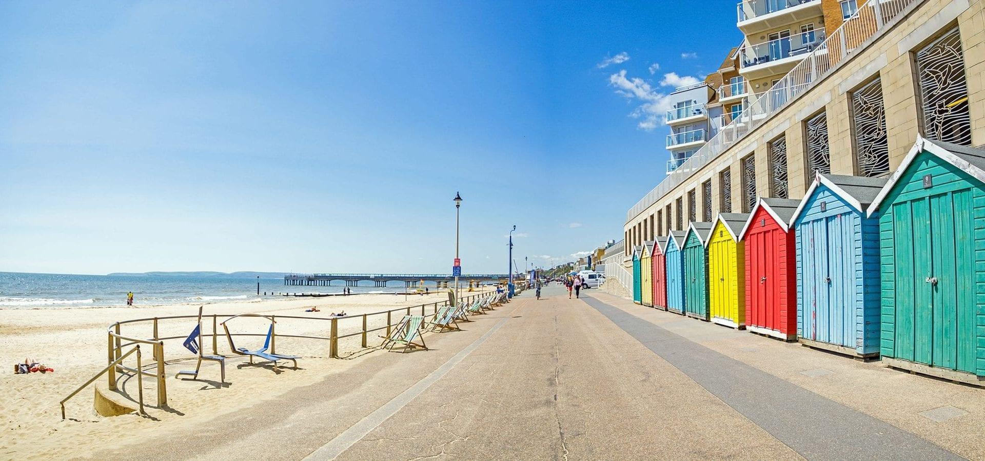 A row of green, blue, yellow and red alternating colourful beach huts on the right, facing the beach and seaside on a sunny day.