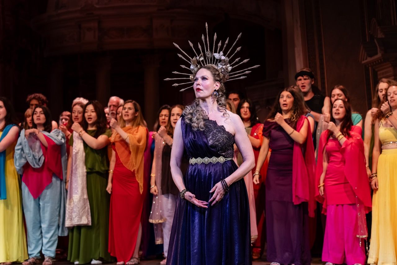 A woman in a dark purple gown and spiked headdress stands centre stage, with a group of singers in colourful costumes behind her.