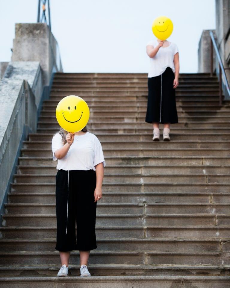 People standing on outdoor steps with yellow smiley balloons covering their faces.