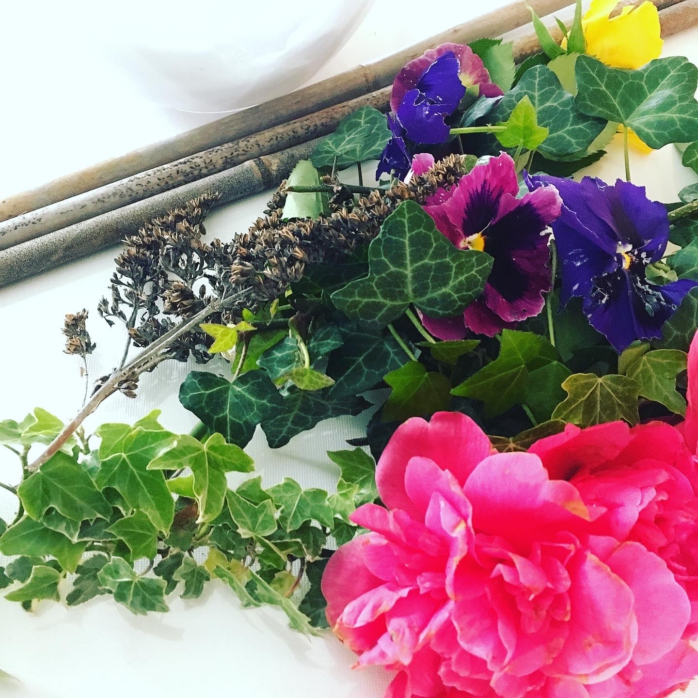 Colourful flowers including pink peonies, purple and yellow pansies with green leaves arranged on white surface beside bamboo canes.