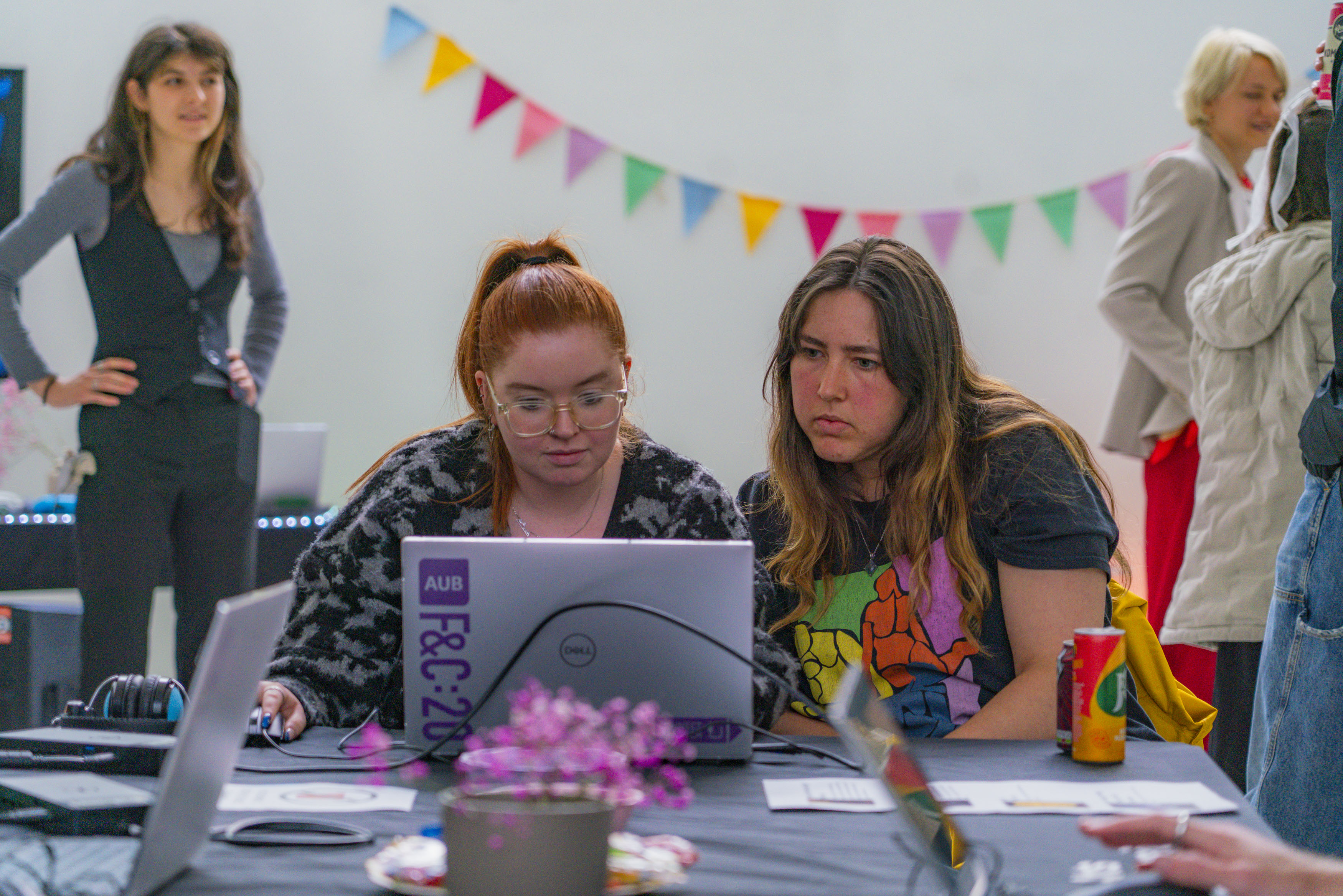 Two students looking at a laptop, one with ginger hair and glasses, the other with long brown hair, colourful pink flowers on table and bunting in the background.