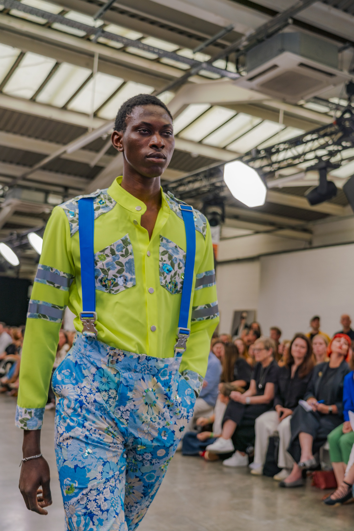 Model wearing hi-vis style garment with colourful blue floral trousers and braces, audience blurred in background.