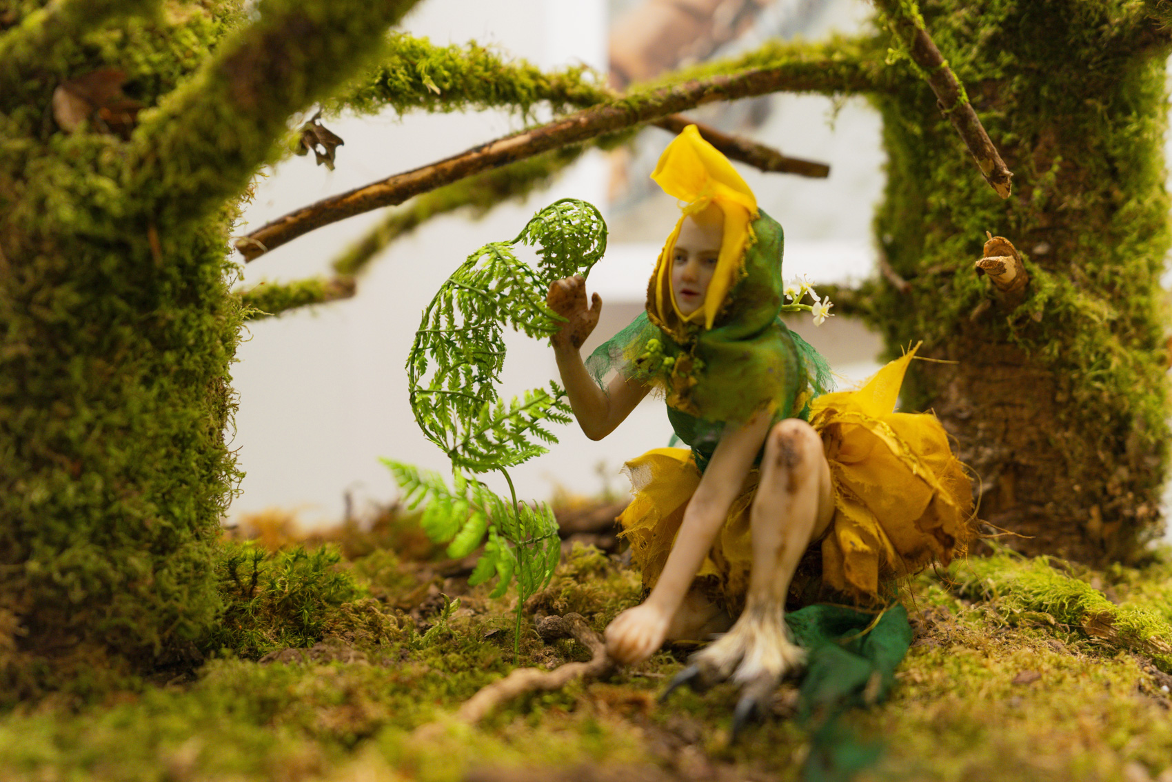 Small model of a fairy-like creature touching a fern, surrounded by green moss and branches.