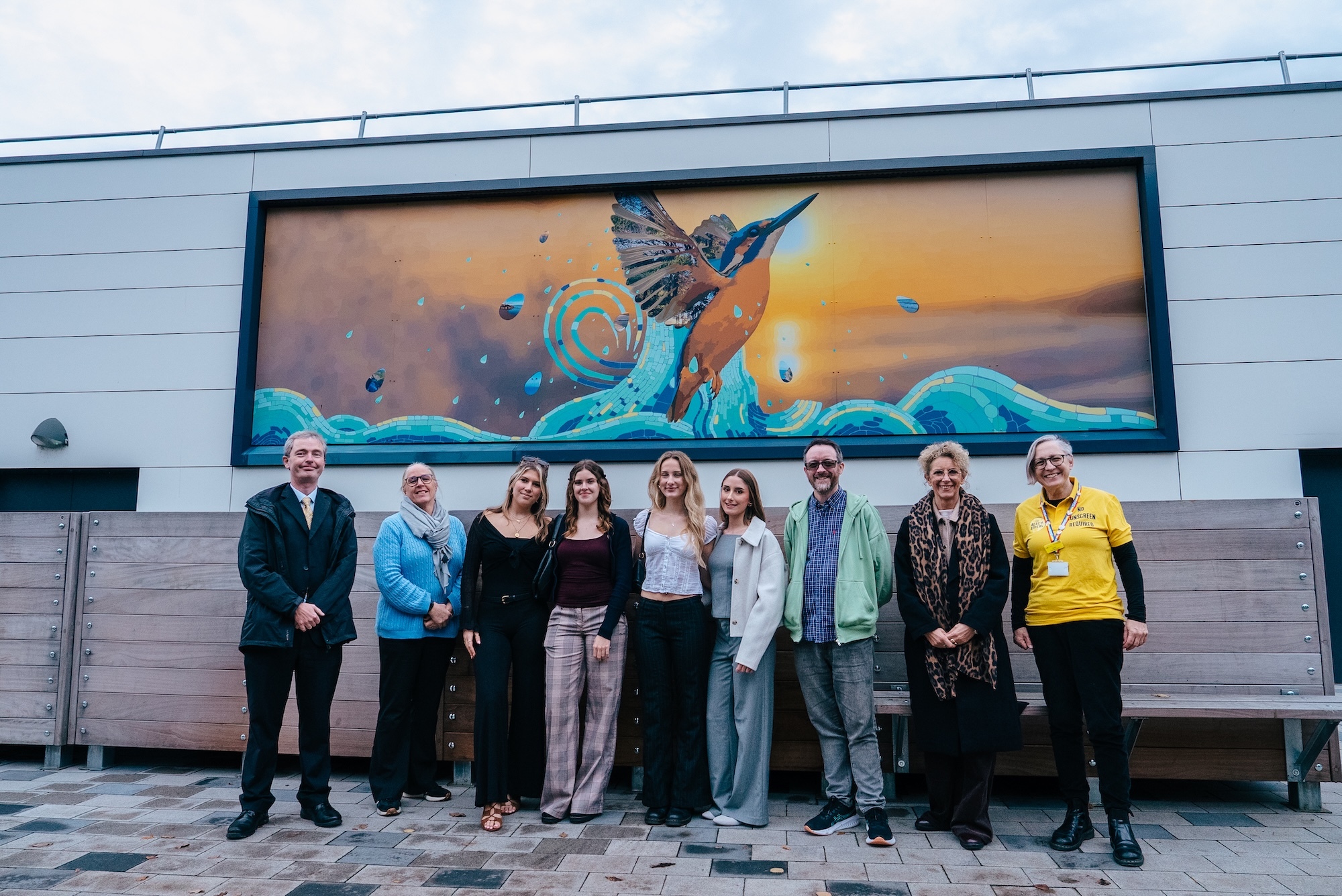 Group of nine people standing in front of large wall mural featuring bird with spread wings, orange sky, and turquoise waves on building exterior.