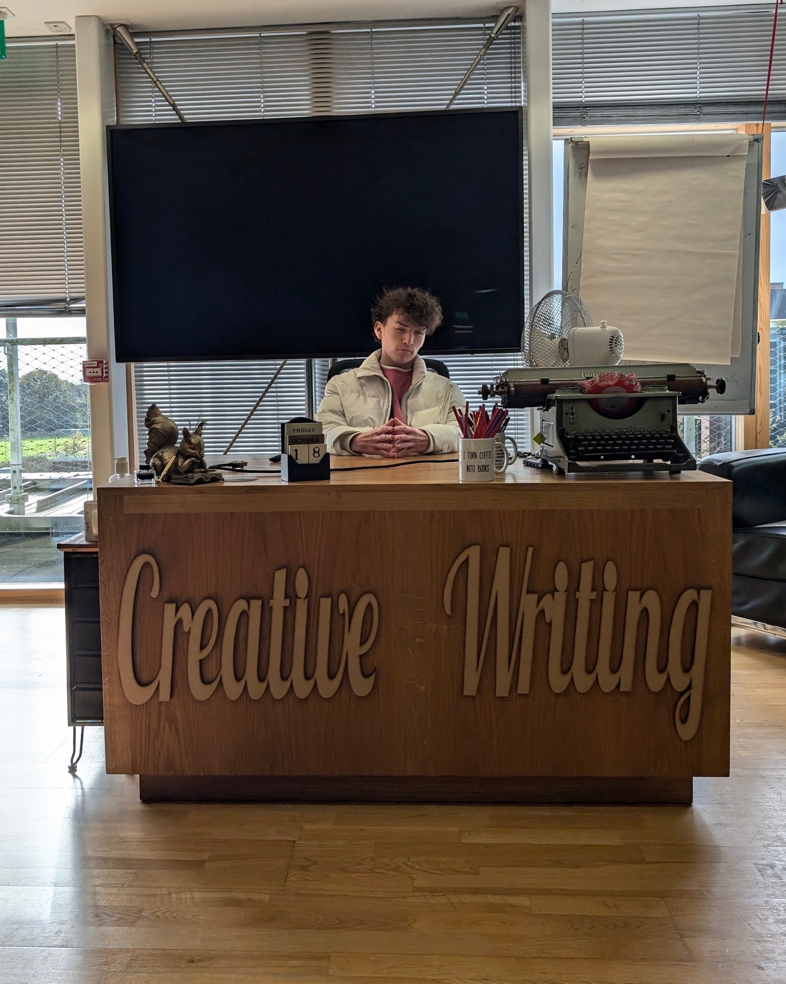 Person in beige jumper sitting behind wooden desk with "Creative Writing" carved text, blackboard and window blinds in background.