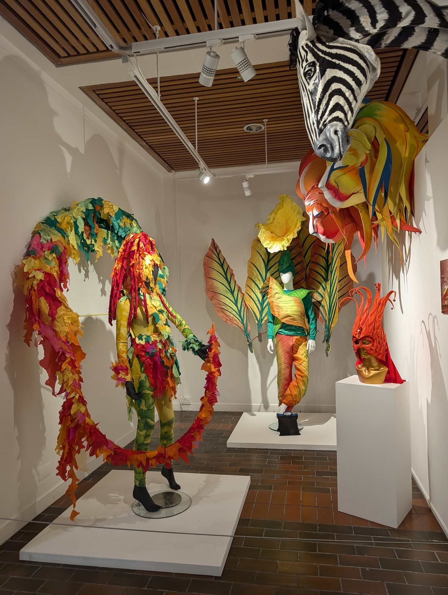 Gallery display with colourful sculptural figures on white plinths. Zebra-striped head with bright feathered costume, curved serpentine form in red, yellow, green plumage.