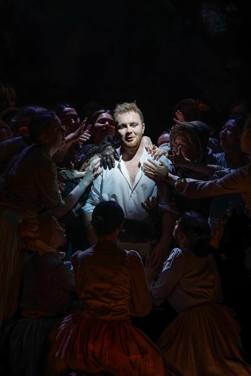 A blond man in a white shirt stands under a spotlight, surrounded by people in dark clothing with their hands raised in a dimly lit stage scene.