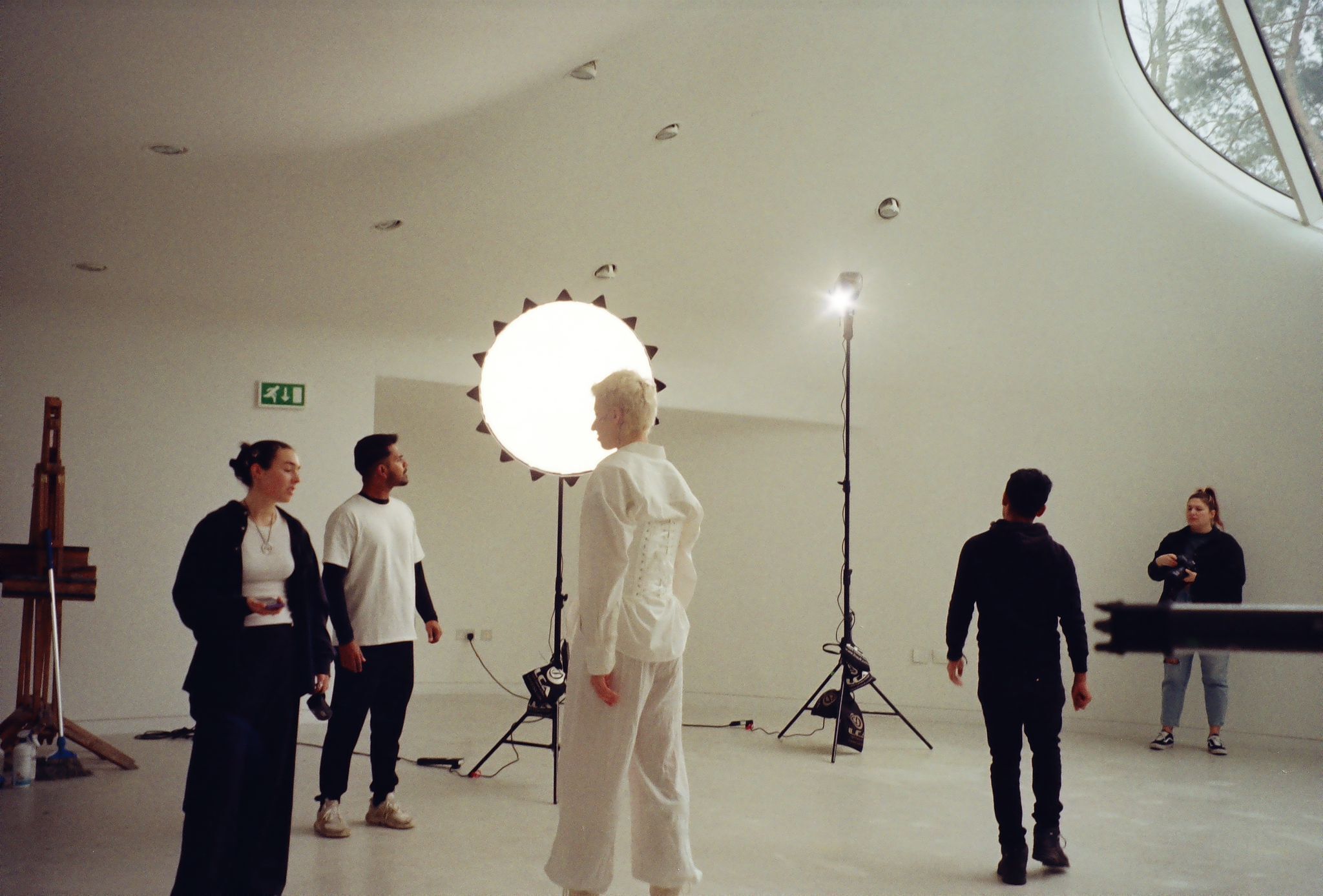 Film crew setting up lighting equipment in modern white interior space with curved ceiling and circular skylight above.