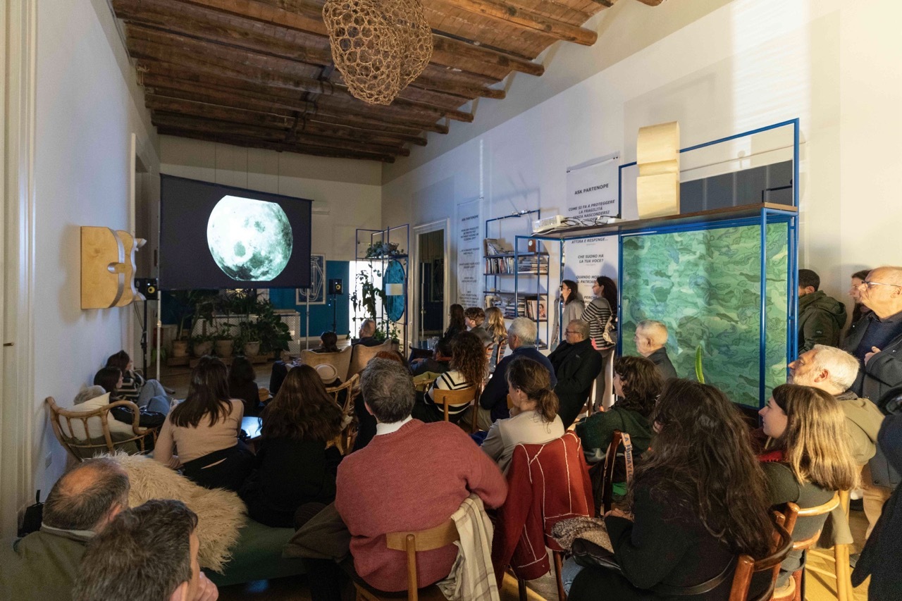 An audience watching a large screen in a room with displays, wooden board ceiling.