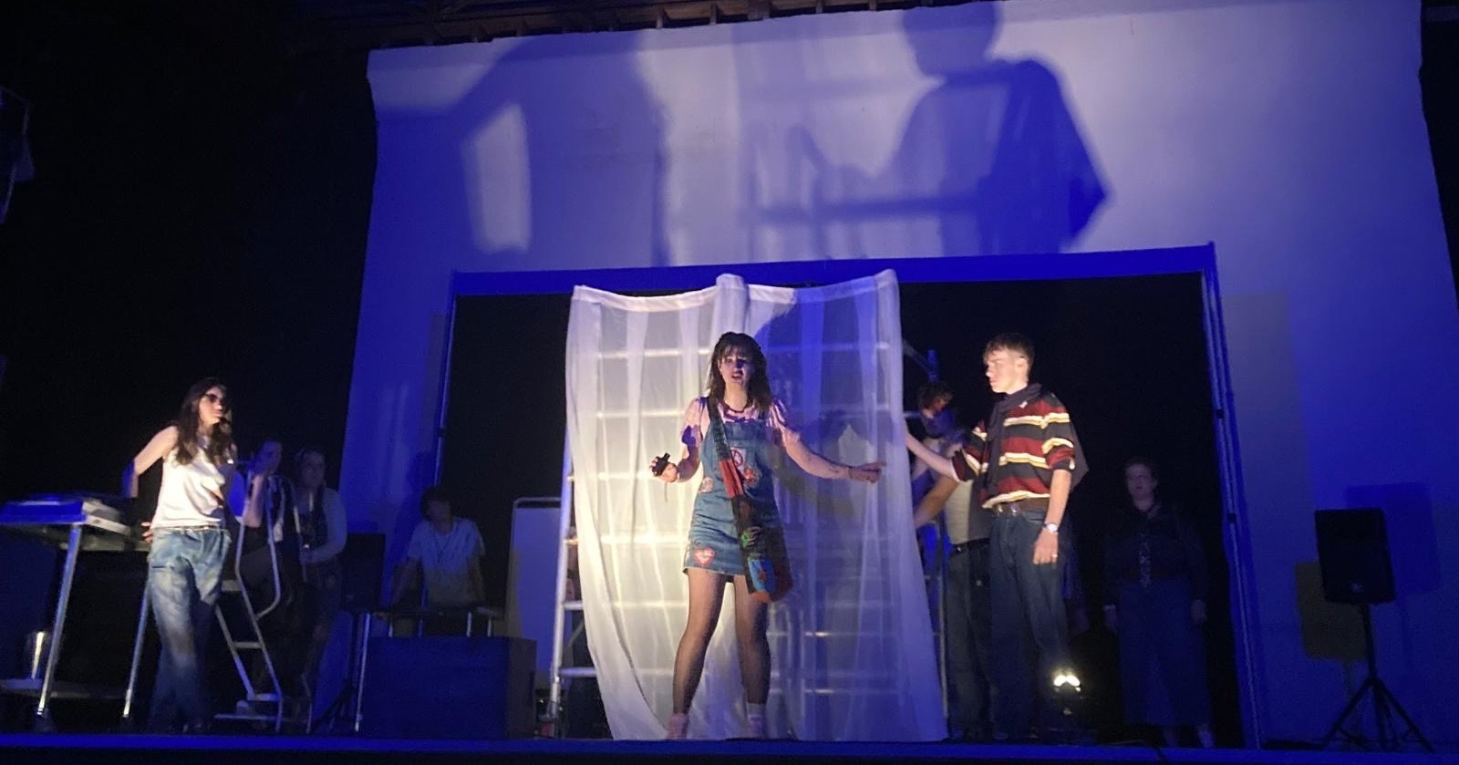 Actors on stage in blue and white dramatic lighting with set design.