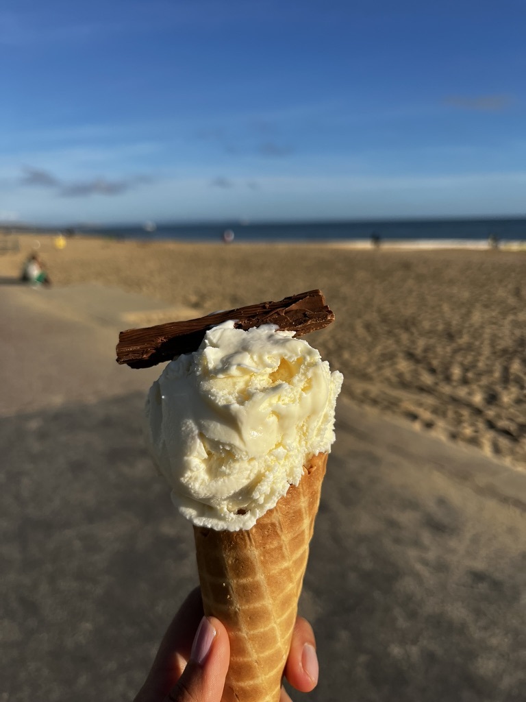 Close-up of a person holding a cone of ice cream with a flake, with Bournemouth Beach in the background