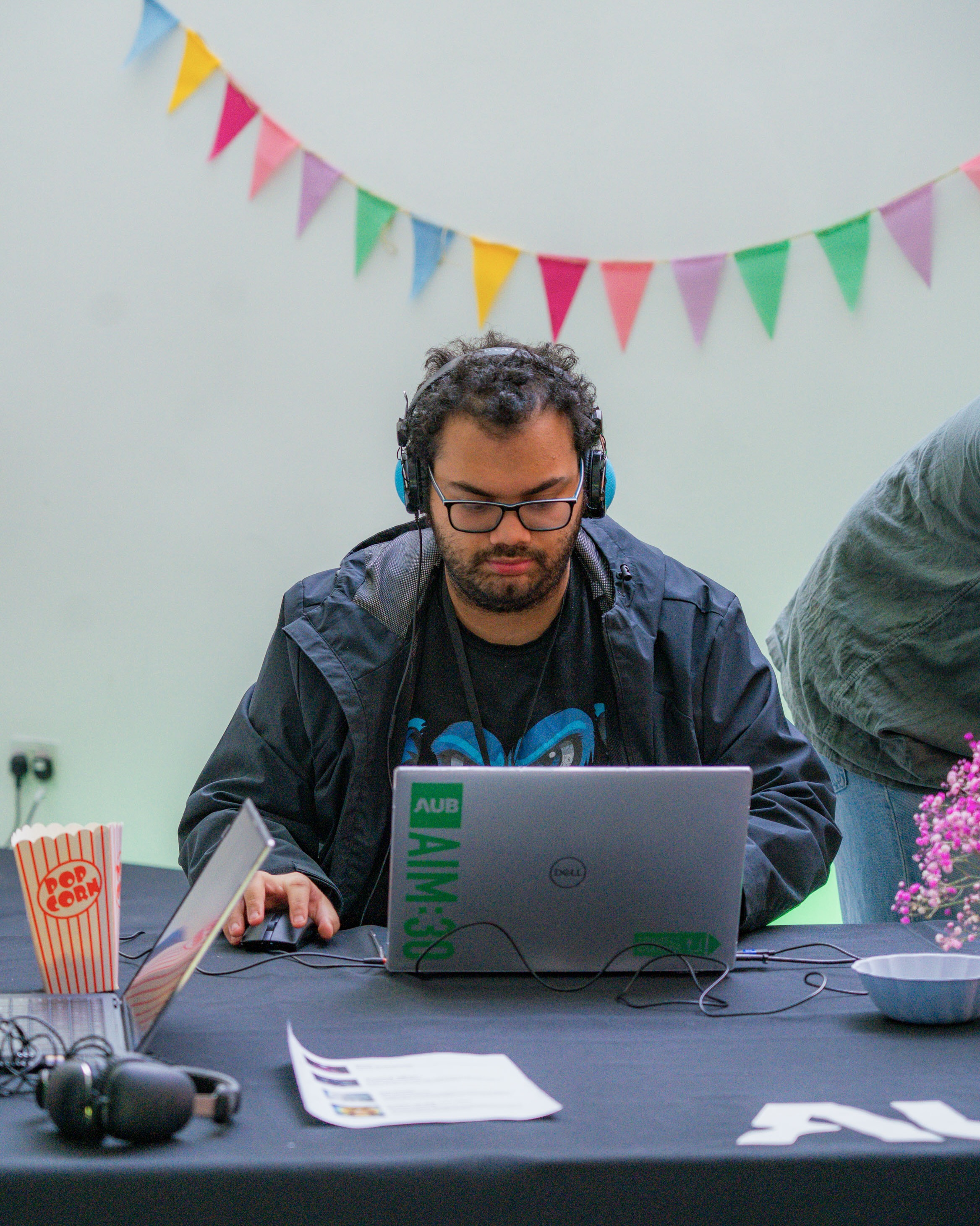 Student wearing blue headphones with glasses and beard interacting with a laptop, colourful bunting in the background.