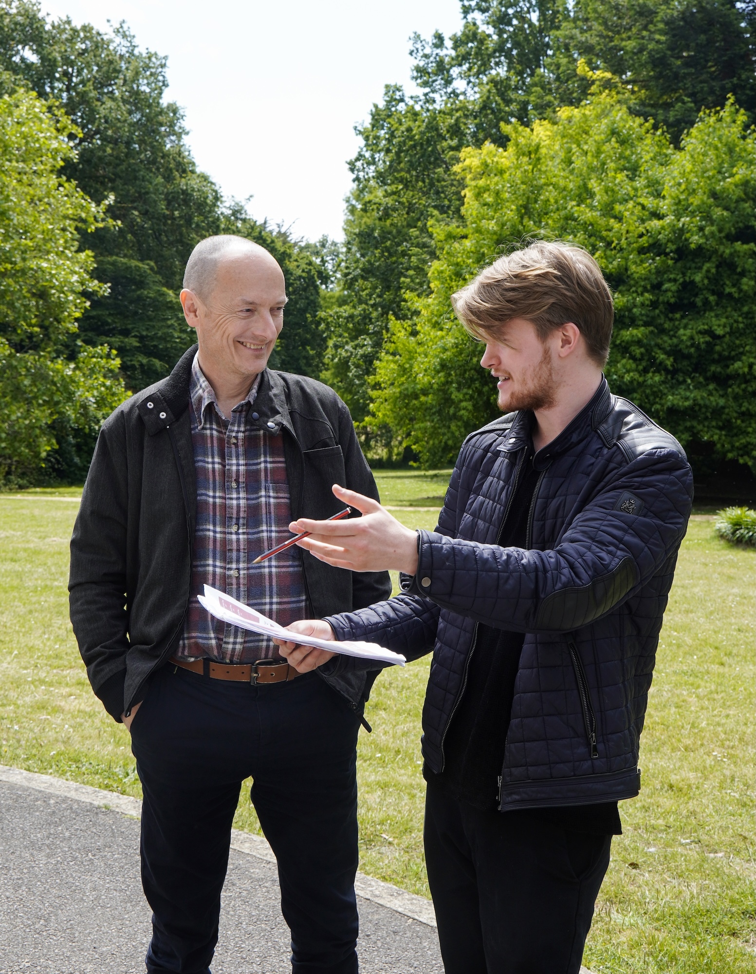 J Rigby and J Hoskins standing outdoors on grass with trees behind them, one bald wearing plaid shirt and jacket, other with brown hair in dark jacket.