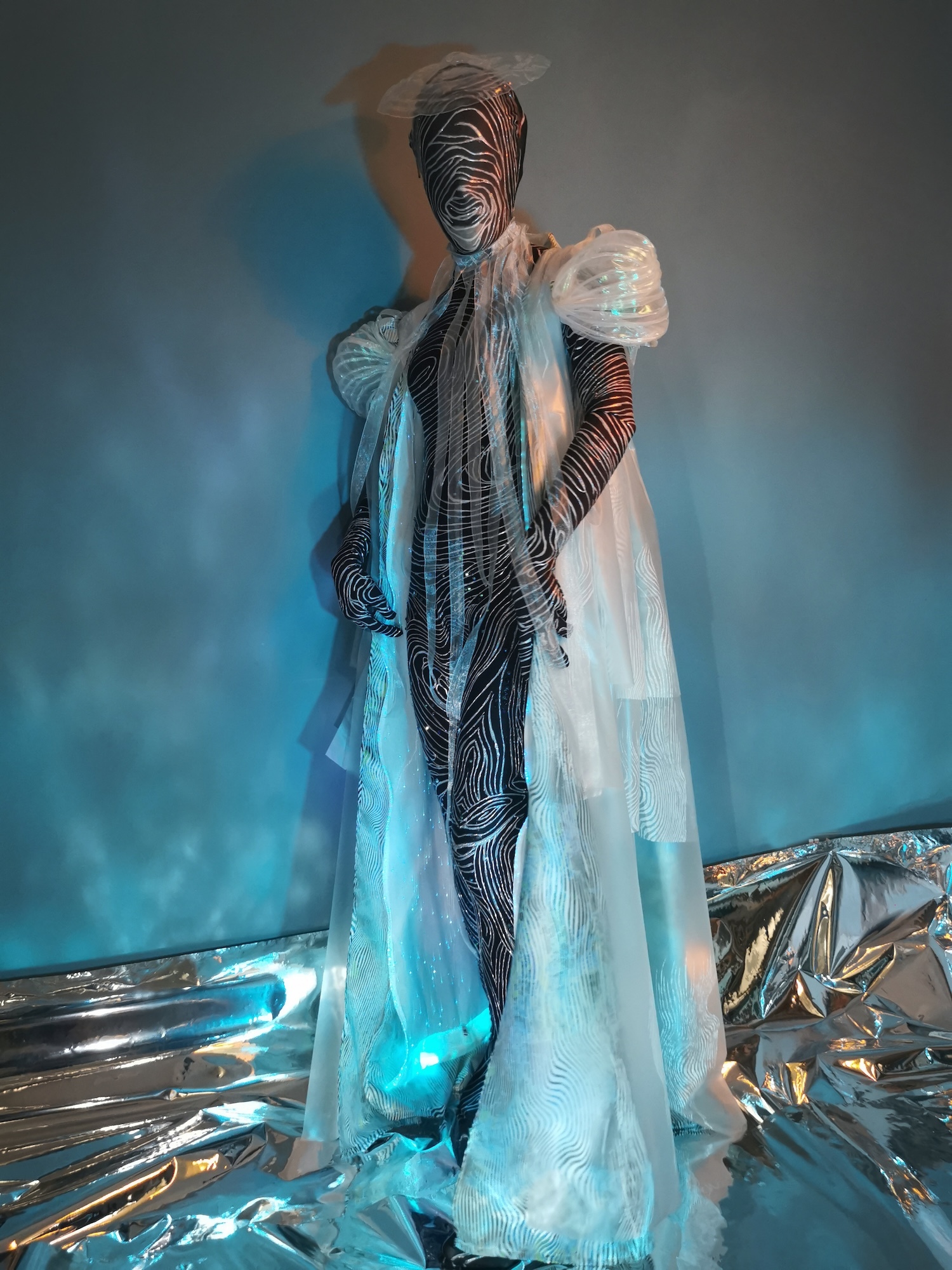 Figure in white flowing fabric with striped pattern overlay, standing on reflective metallic surface with blue lighting against grey backdrop.