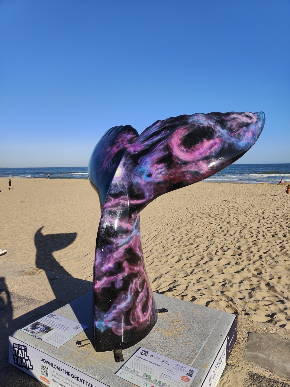 Dark purple and pink galaxy-style mermaid tail sculpture at the beach, blue sky and sunny.
