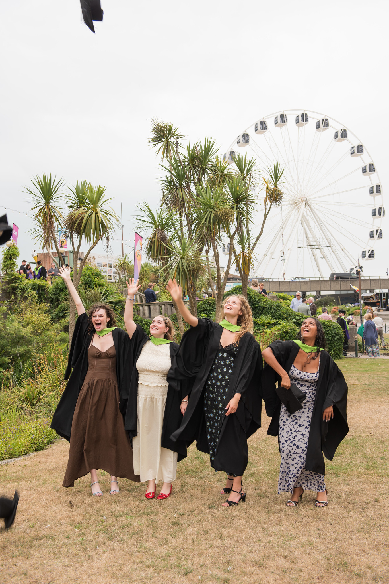 Four graduates in black gowns and green sashes throwing graduation hats into air, with palm trees and white ferris wheel in background.