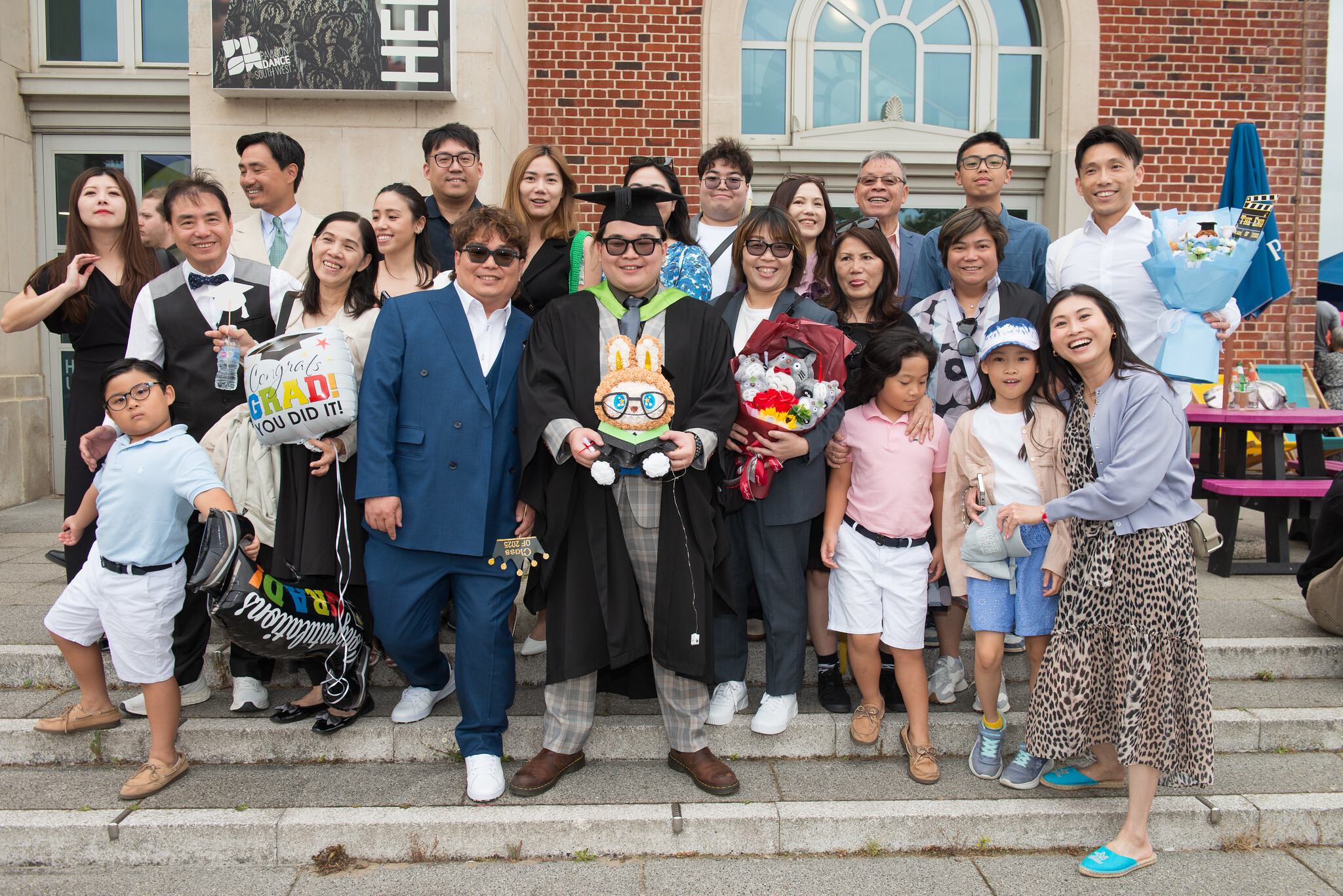 Large group of people posing on brick building steps, one wearing a graduation gown, including suits, formal wear, and outfits with props.