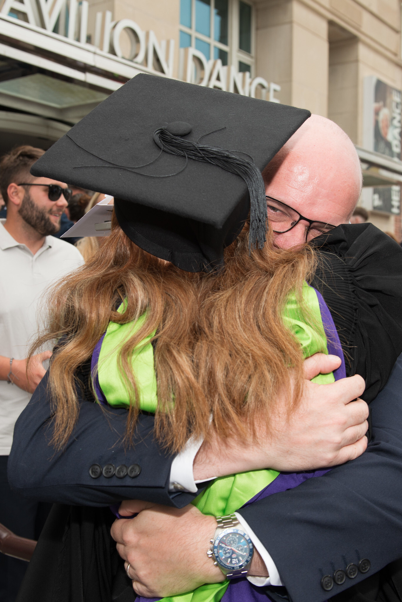 Graduate in black cap and gown embracing person in bright green top outside building.
