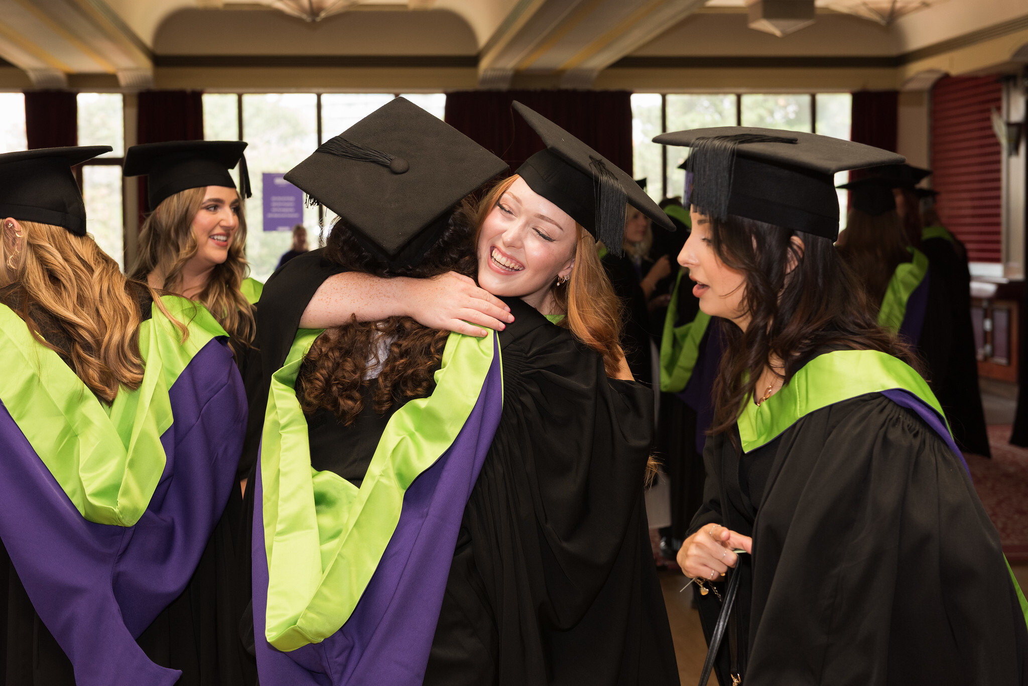 Graduates in black caps and gowns with green and purple trim embrace and celebrate indoors near large windows.