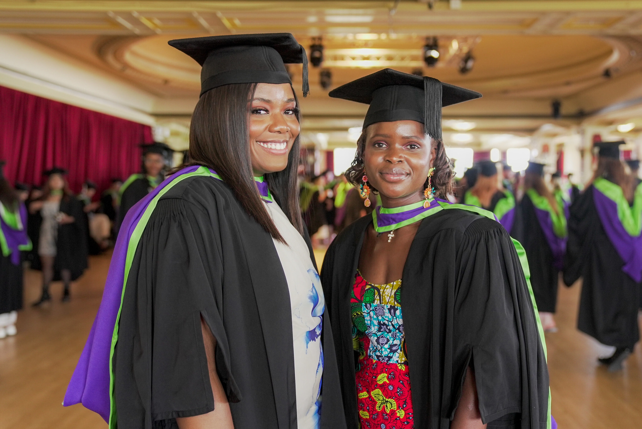 Two women in black graduation gowns with purple hoods and mortarboards smile at graduation ceremony in ornate hall with red curtains.