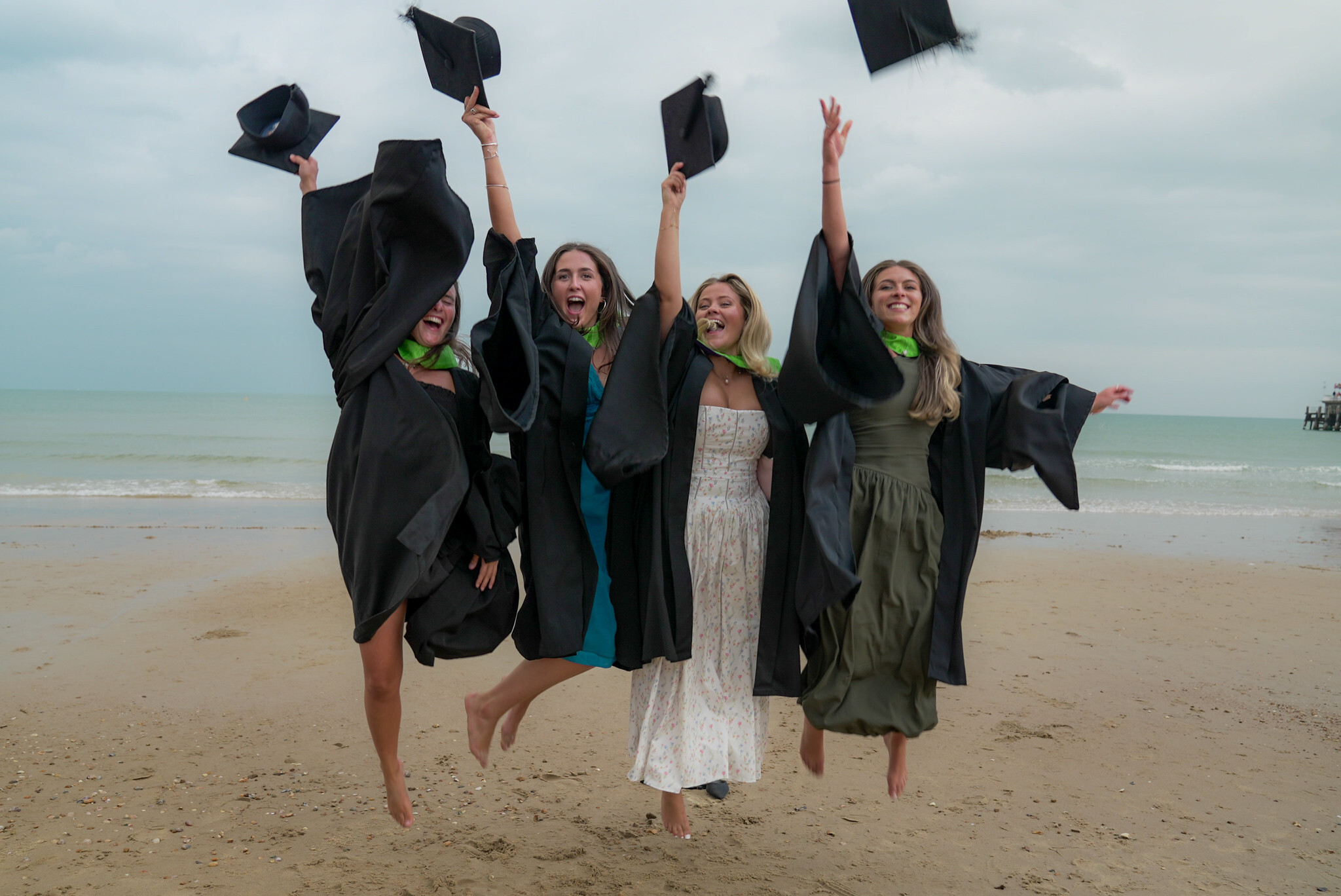 Four graduates in black gowns throwing graduation hats in air on beach, arms raised, overcast sky and sea in background.