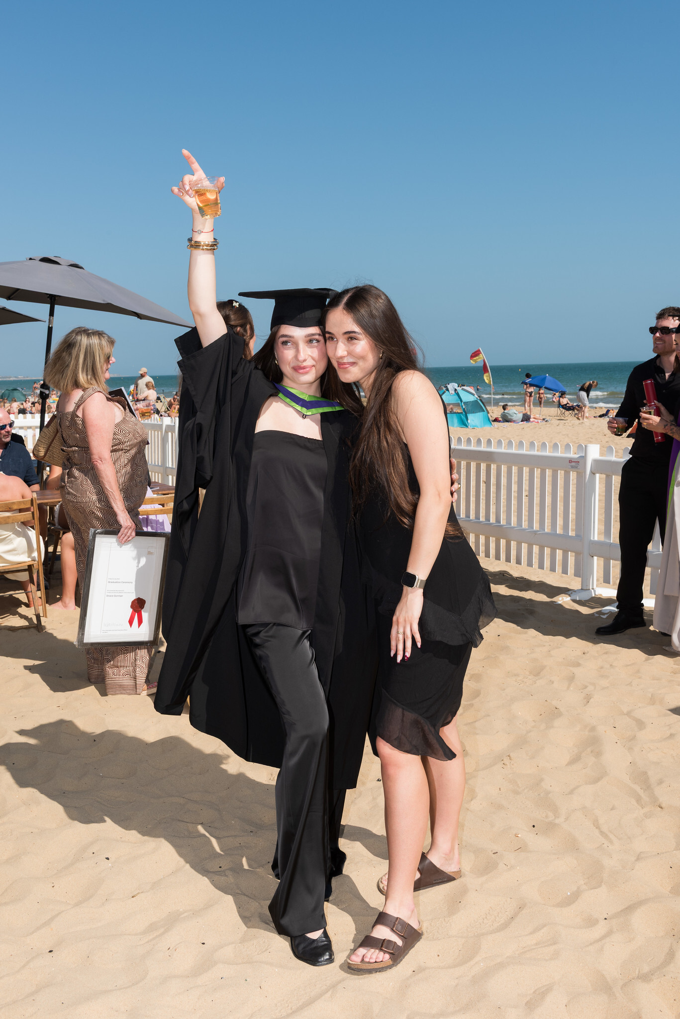 Graduate in black cap and gown celebrating with friend on beach, raising arm in victory pose, blue sky and white fence behind.