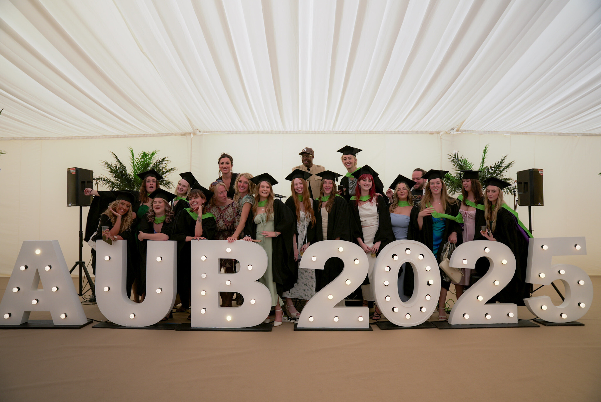 Group of graduates in caps and gowns posing behind large white "AUB 2025" letters with light bulbs, white draped ceiling above.