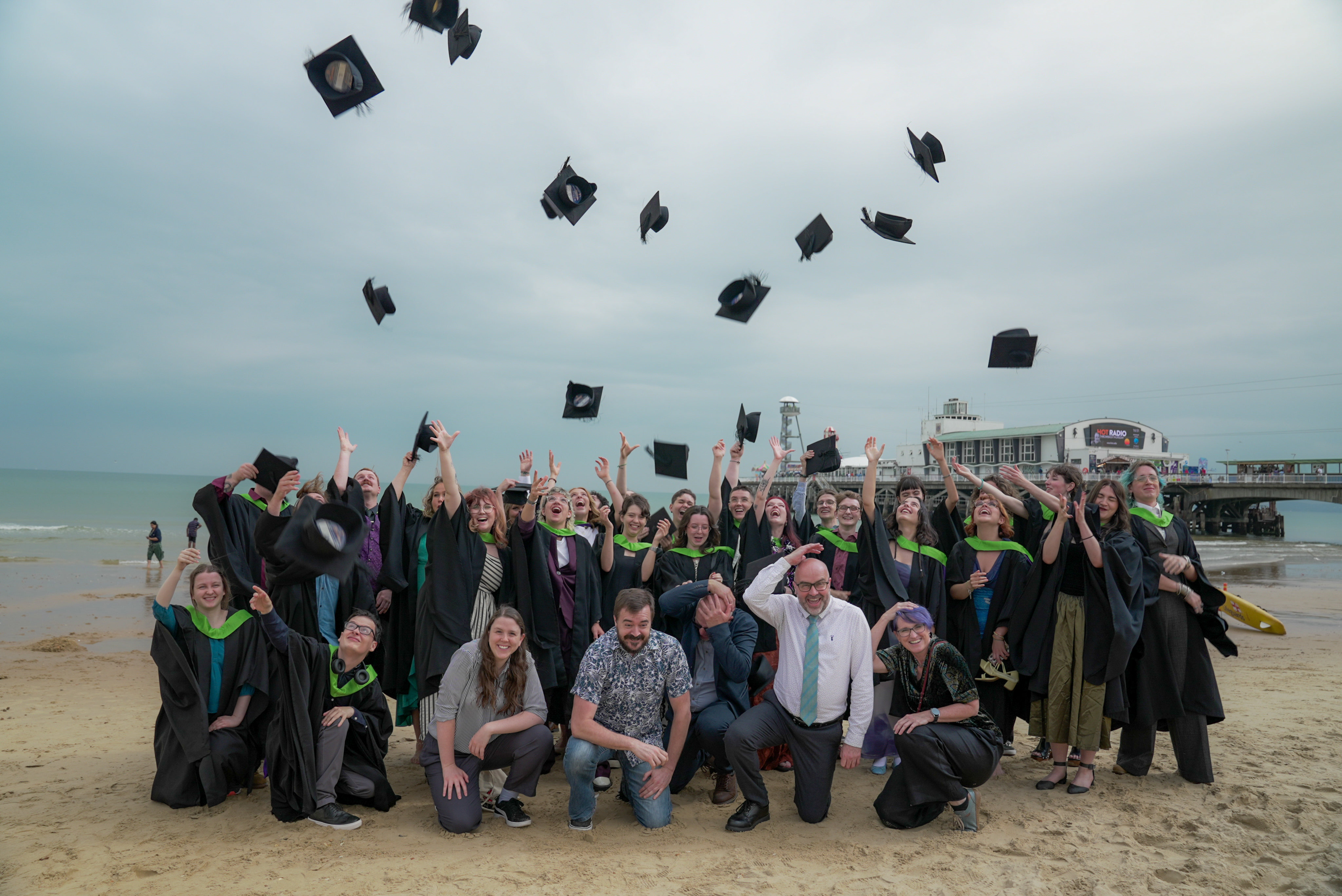 Graduation ceremony on beach with students in black gowns throwing mortarboards into grey sky, pier visible in background.
