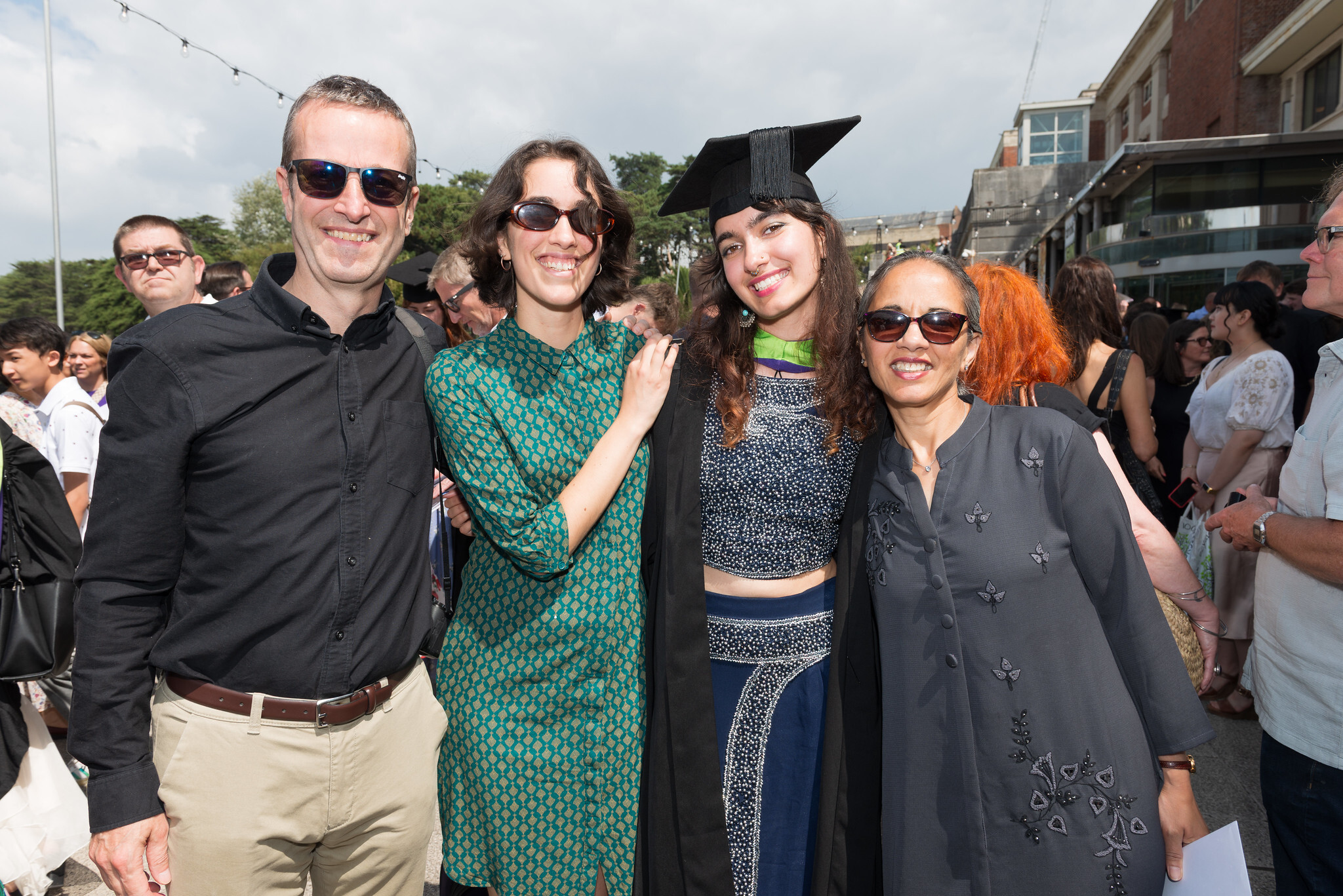 Four people posing together at outdoor graduation event; man in black shirt, woman in green dress, graduate in cap and gown, woman in grey top.