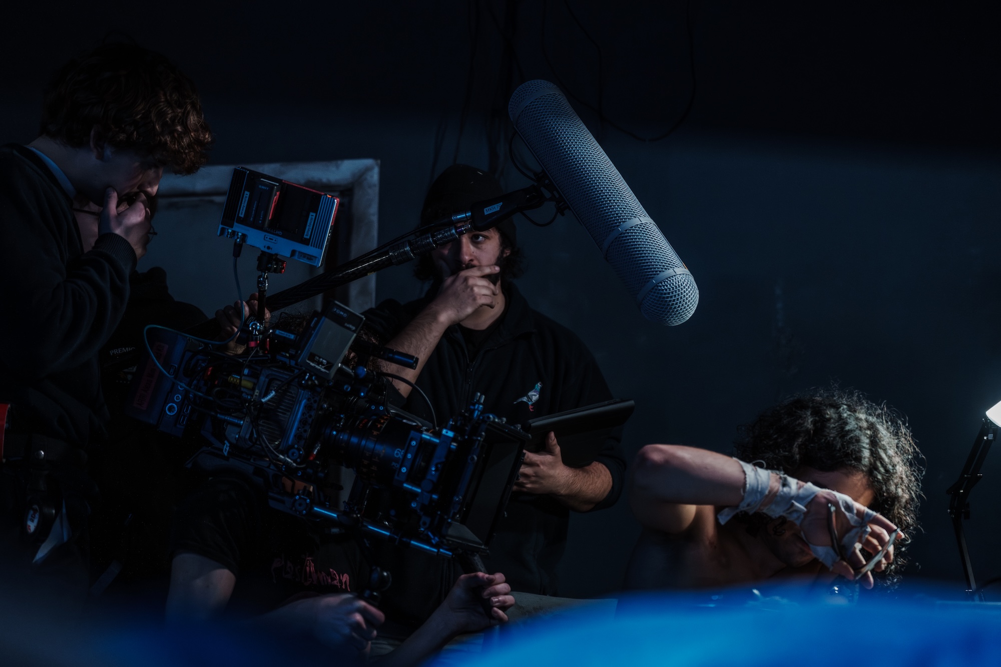 Crew members work around a film camera in a dark studio, with a large boom microphone hanging overhead.