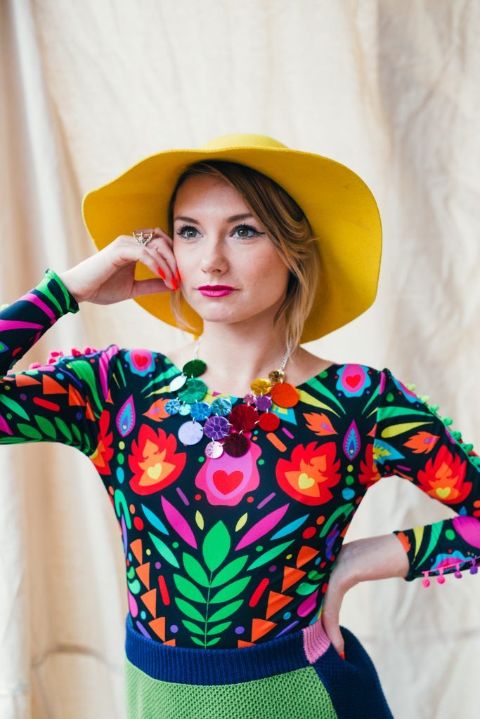 Headshot of alumna T Burnett, who is standing wearing a colourful shirt and bright yellow hat.