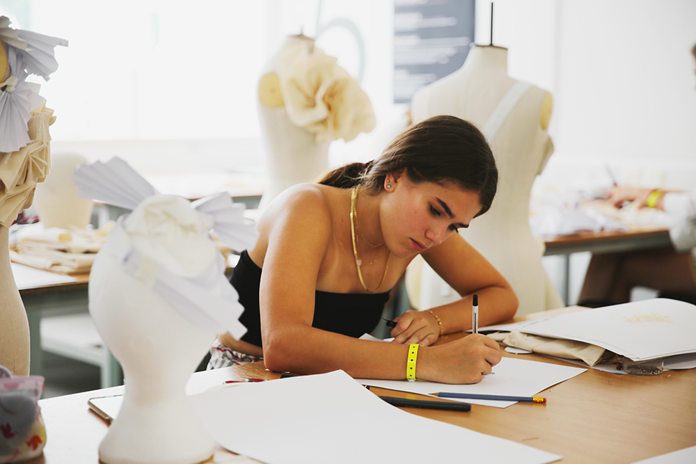 Woman in black top writing at desk in fashion design studio with dress forms, yellow flowers, and fabric samples visible.