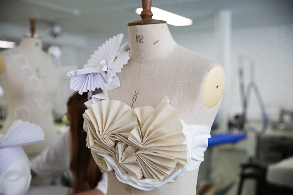 Cream dress form displaying white pleated fabric pieces and paper fan decorations in fashion design studio.