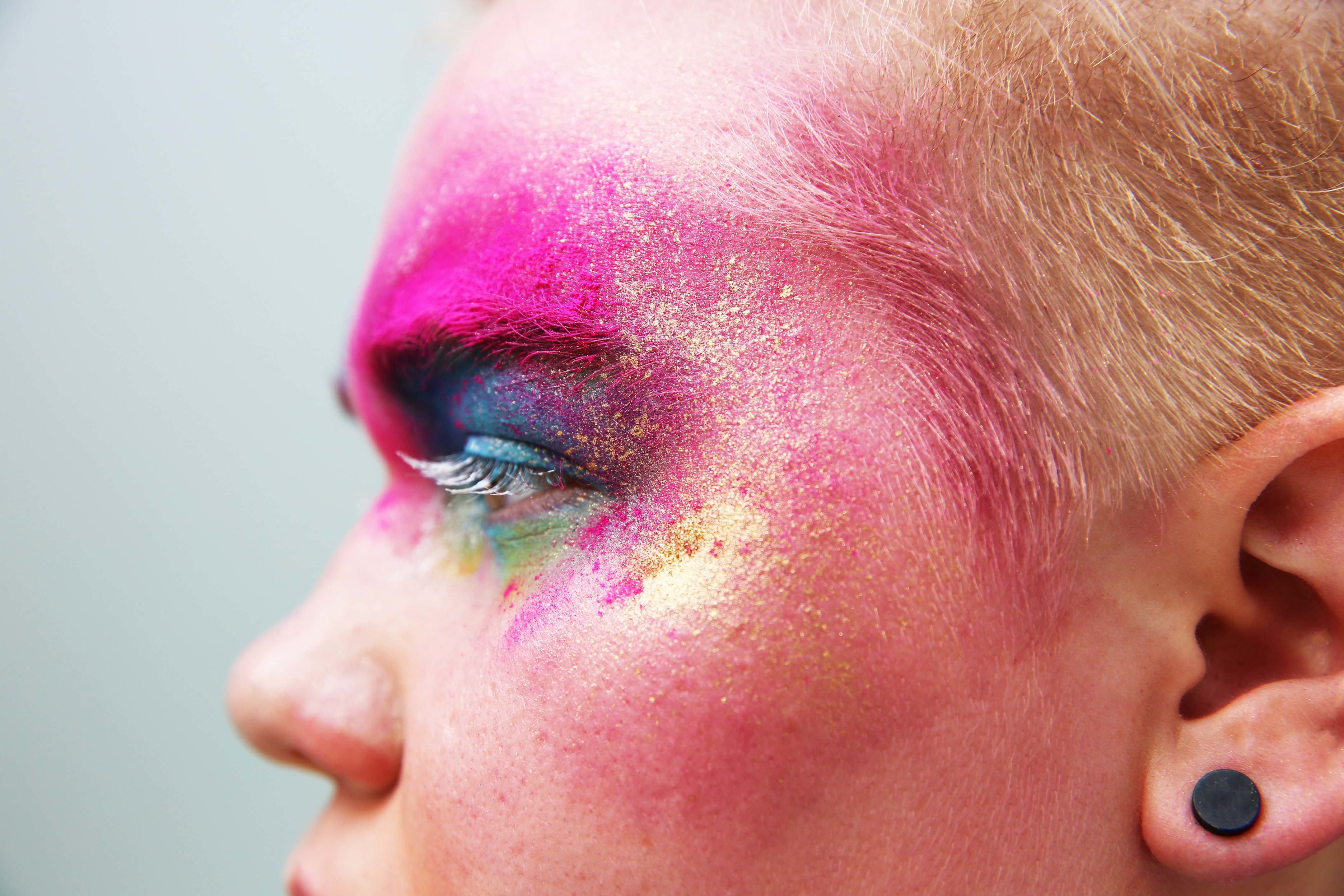 Profile view of face with vibrant pink, purple, and green powder makeup around eye area, blonde hair, black ear plug visible.