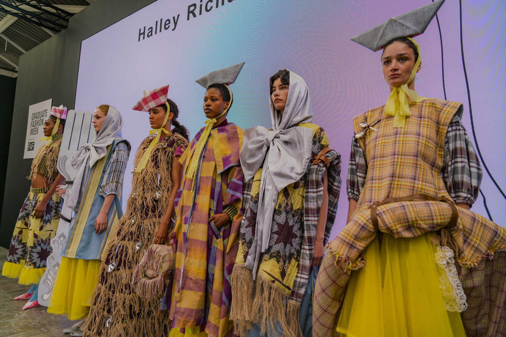 Line-up of models wearing vibrant yellow-patterned garments, unique folded headpieces, bows and textures, white screen background.