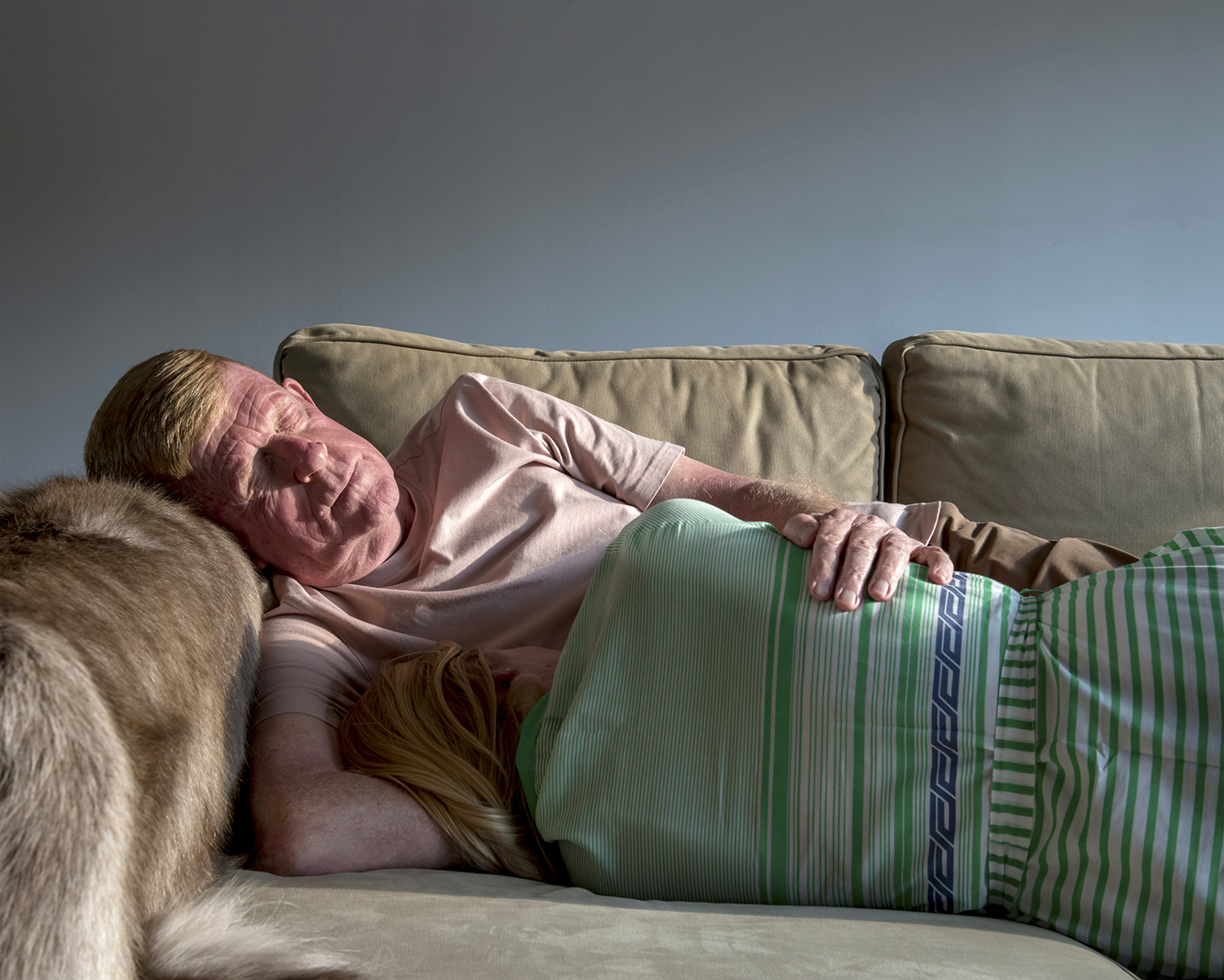 A man lies sleeping on a sofa. A girl lies with her back to the camera and her face buried in the man's chest.