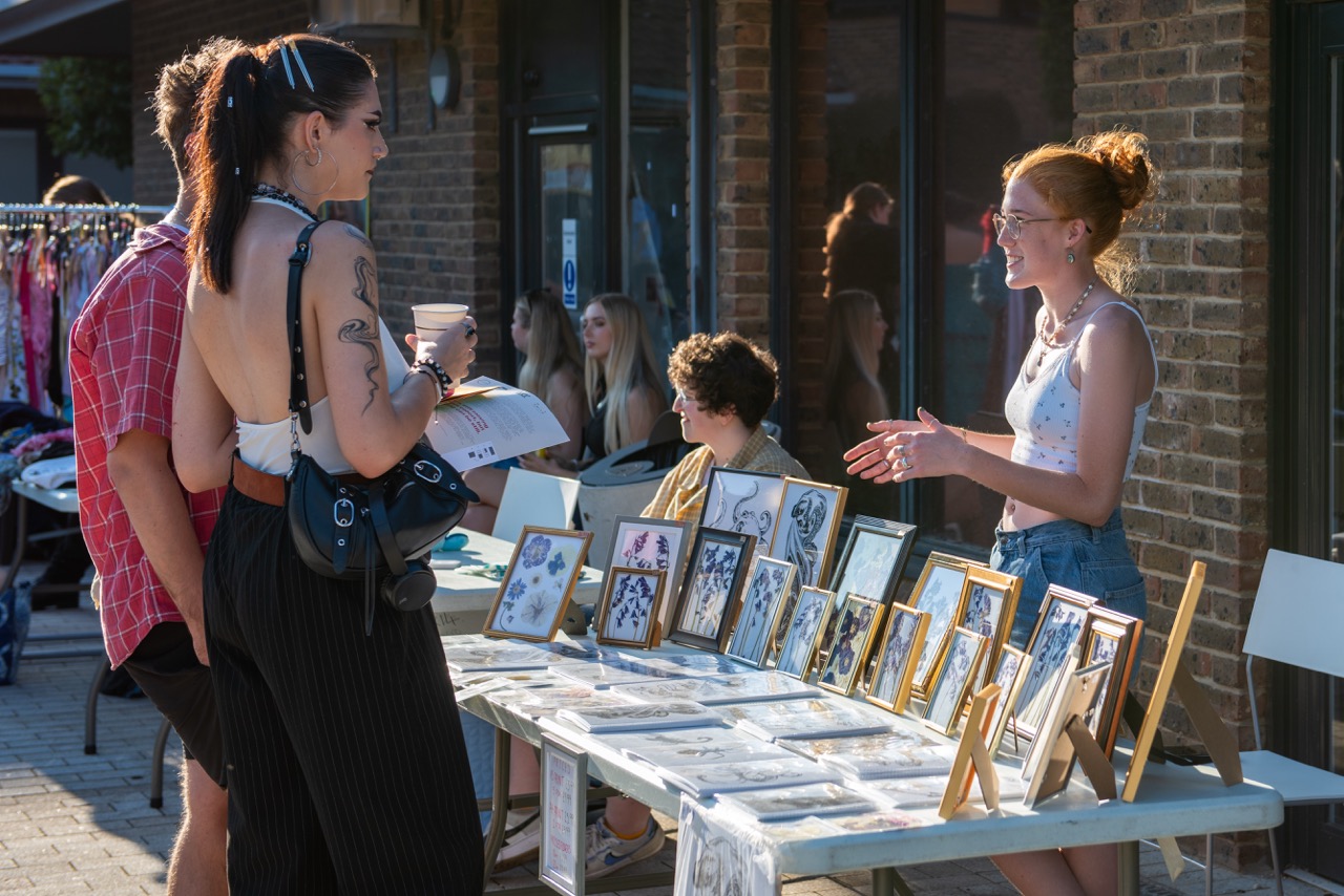 Students stood at a stall with illustrator talking to them, framed floral artwork on the table, outside in sun.