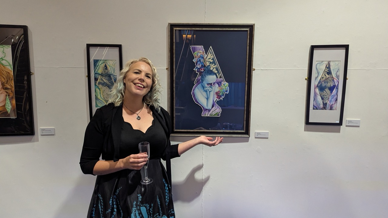 A smiling blonde woman in a black dress stands in a gallery and holds a glass, with framed abstract artworks displayed on a white wall behind her.