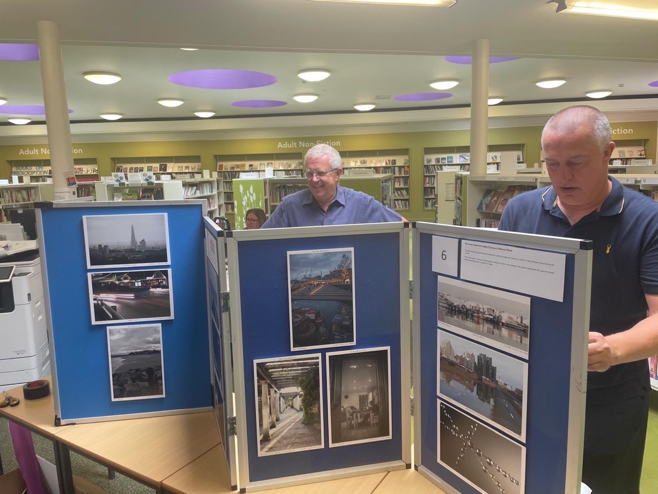 Two men examining blue display boards with mounted photographs in library setting with bookshelves.
