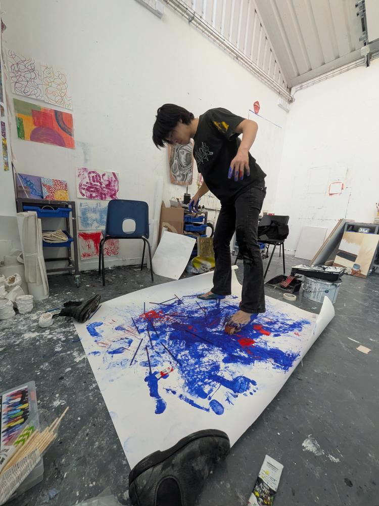 Seiji Suzuki in an art studio standing on paper, blue and red paint splattered which he is drawing with bare feet. Chairs and artwork behind.