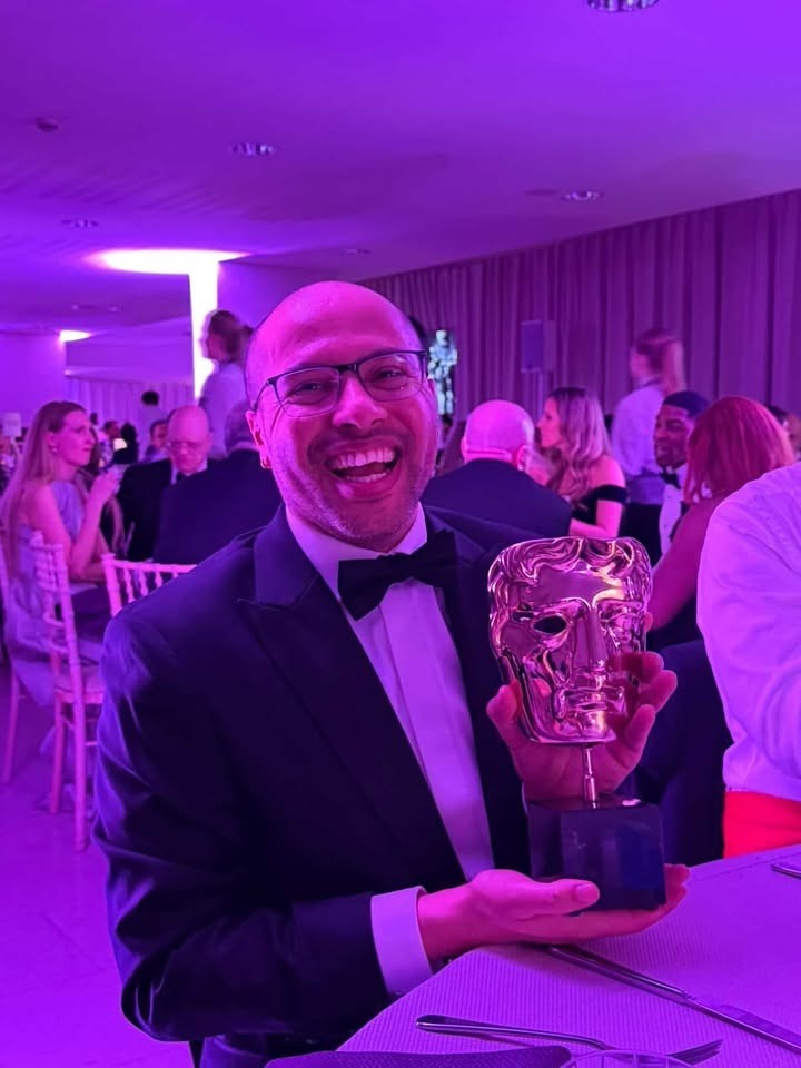 D Atta holding BAFTA award, smiling at camera in purple-lit event space with guests in background.