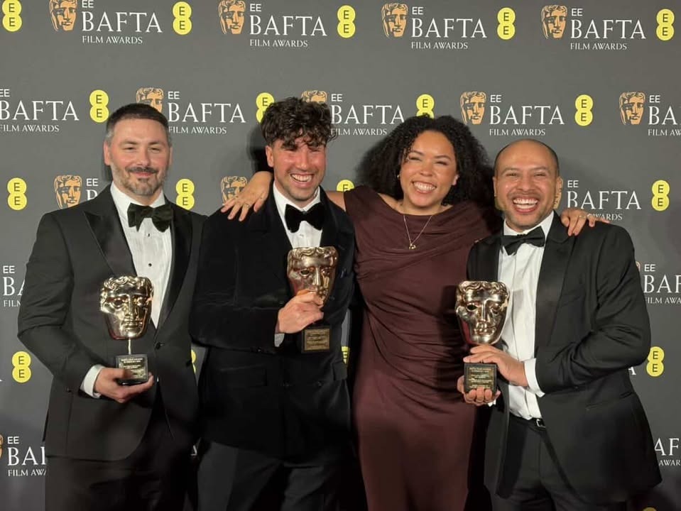 Four people in formal attire holding BAFTA trophies at awards ceremony, three men in black tuxedos and one woman in brown dress.
